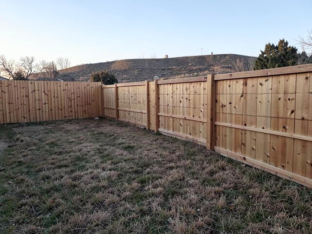 New cedar privacy fence panels along a modest backyard with dry grass and a low ridge behind