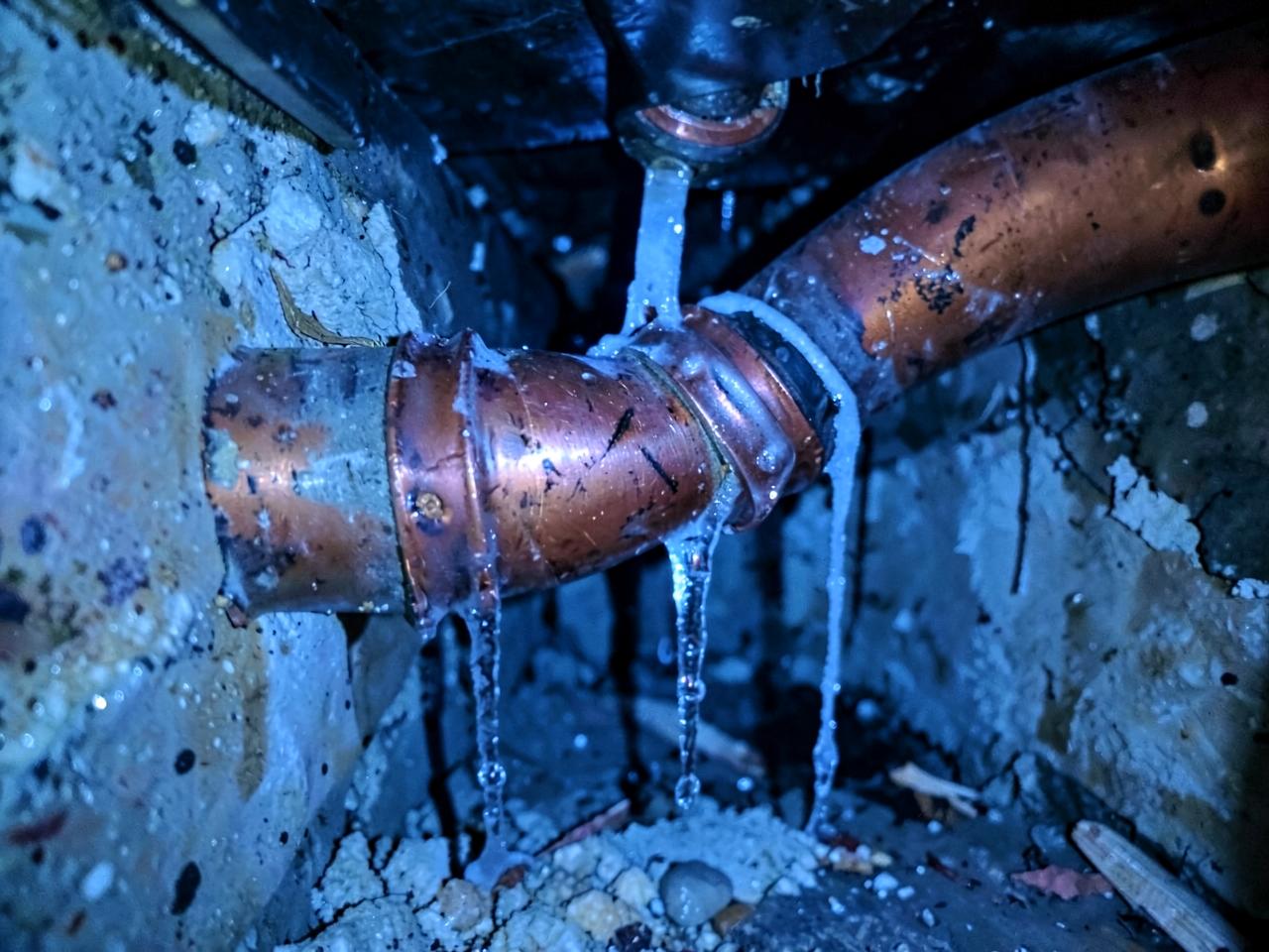 Burst copper pipe with icicles hanging inside a dark crawlspace wall cavity in cold blue light