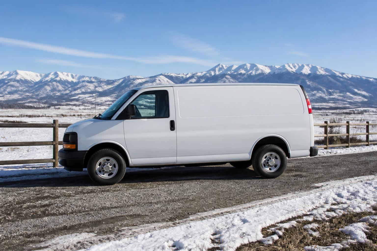 Plain white cargo van with blank panels parked on a snowy gravel farm lane beside a wood rail fence, winter fields and mountains behind