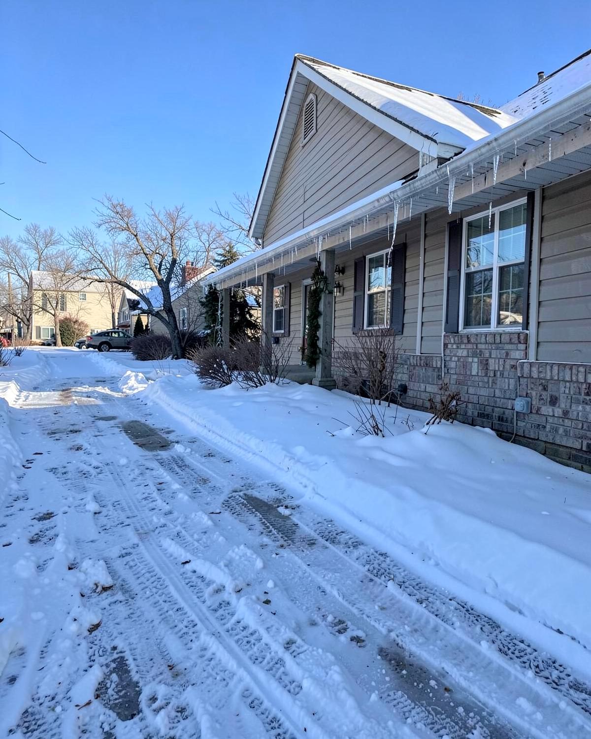 Snow-covered driveway and shoveled path beside a suburban home with icicles along the gutter