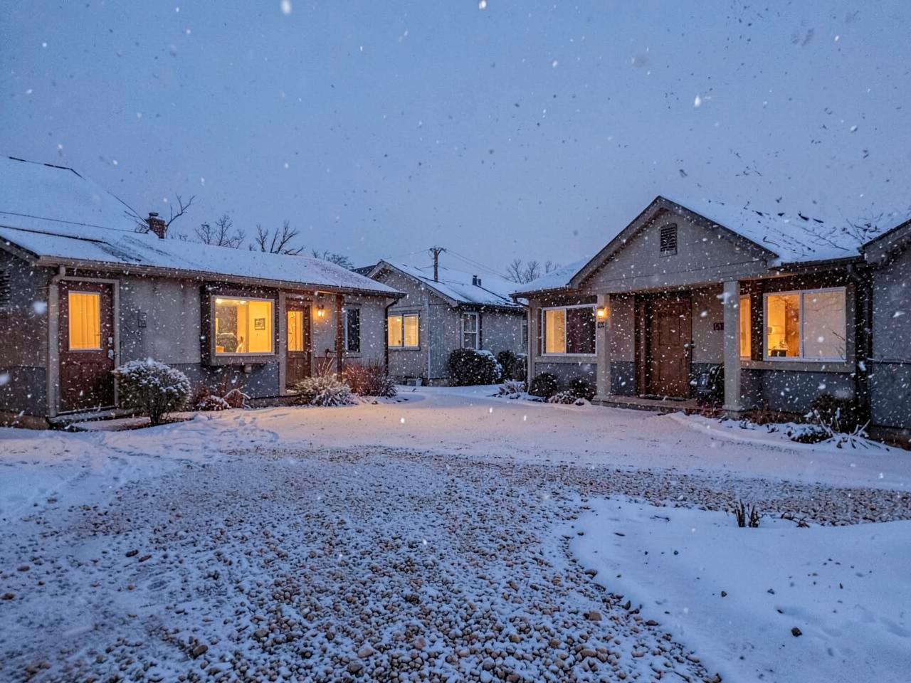 Snow falling at dusk over single-story homes with lit windows and gravel yards under fresh snow