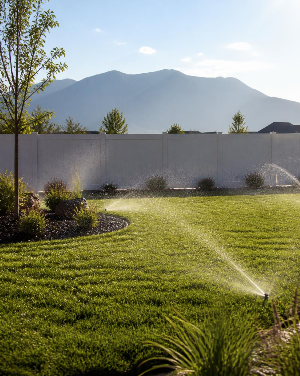 Sprinklers running over a fresh green lawn in a fenced new suburban backyard with a hazy mountain ridge behind