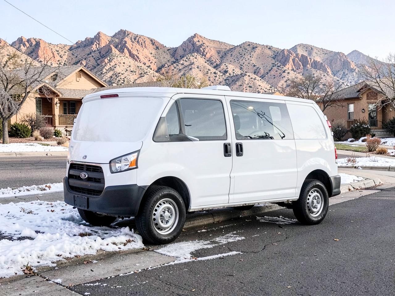 Plain white contractor van parked at a snowy curb on a residential street below rocky desert mountains