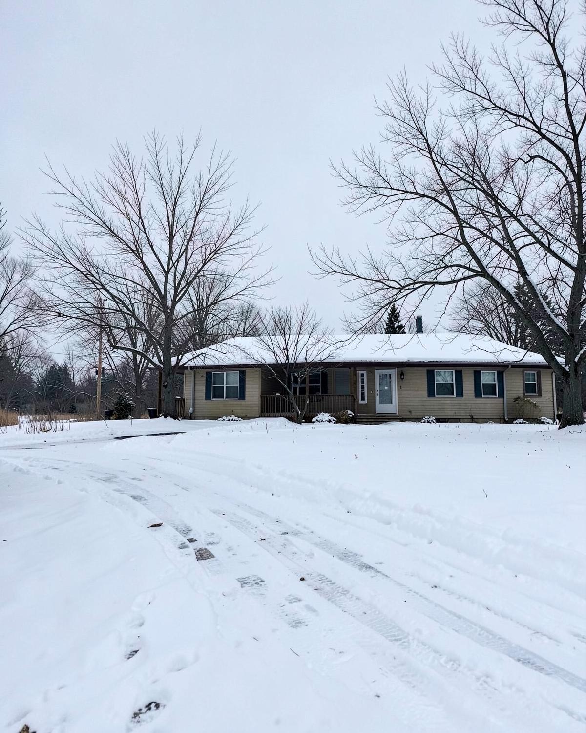 Snow covered rural driveway and bare trees beside a modest single story home in winter
