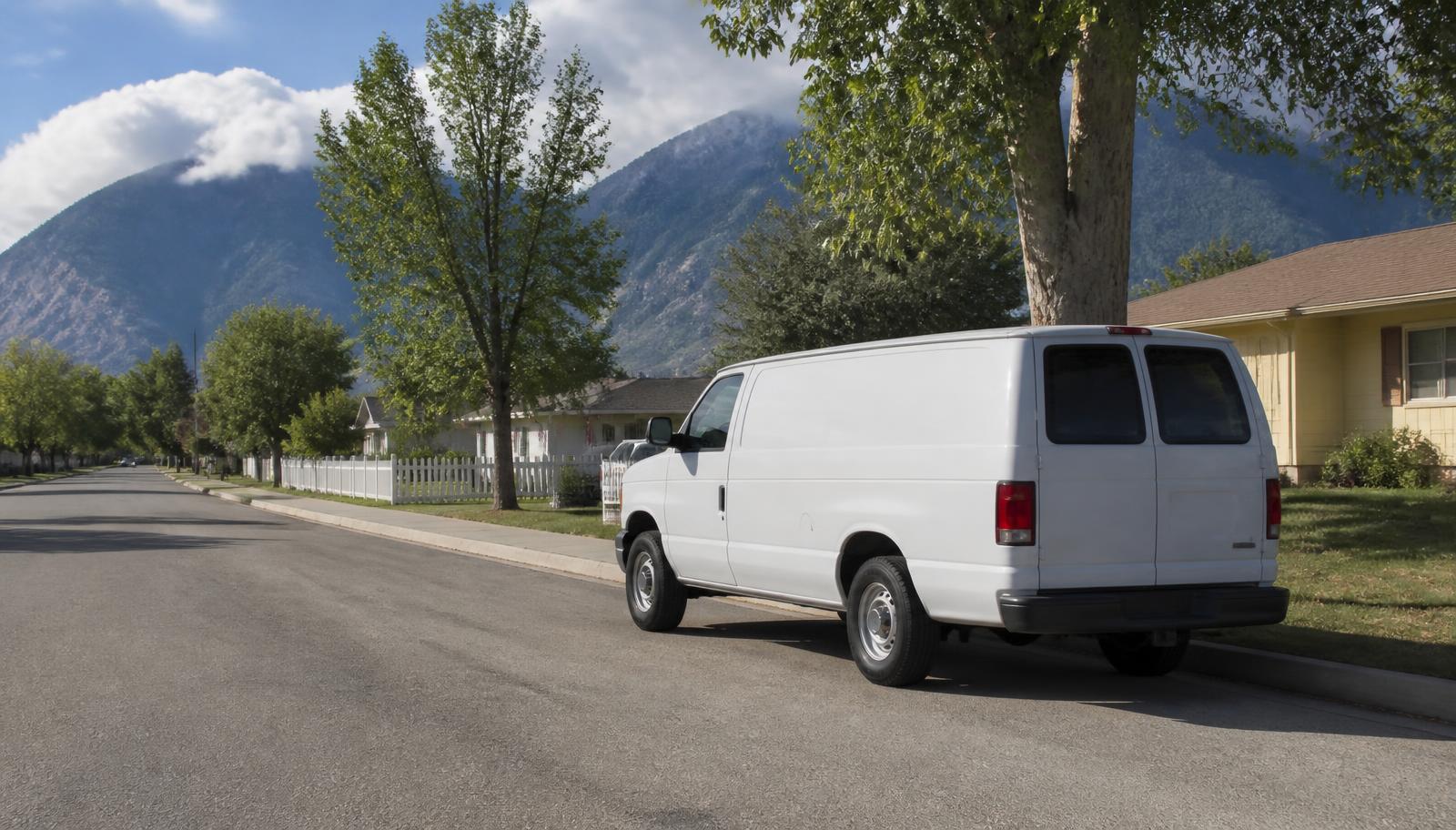 Unmarked white work van parked at a suburban curb with a long mountain ridge visible in the distance