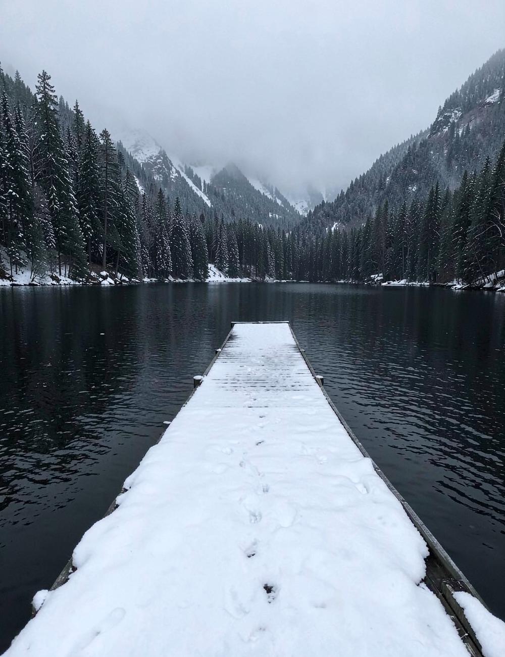 Wooden lake dock covered in fresh snow with misty forested mountains across dark winter water