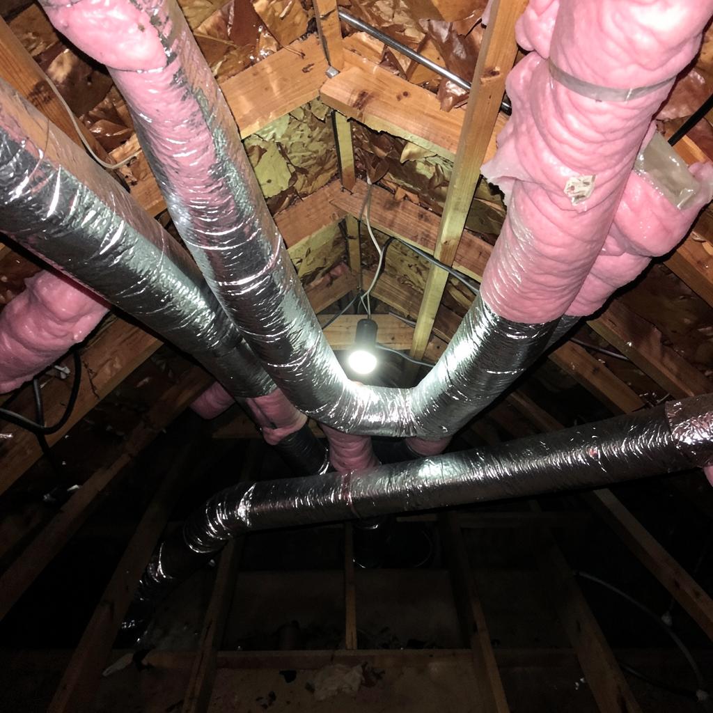 Insulated flexible ductwork runs across joists in a hot residential attic lit by a single work light