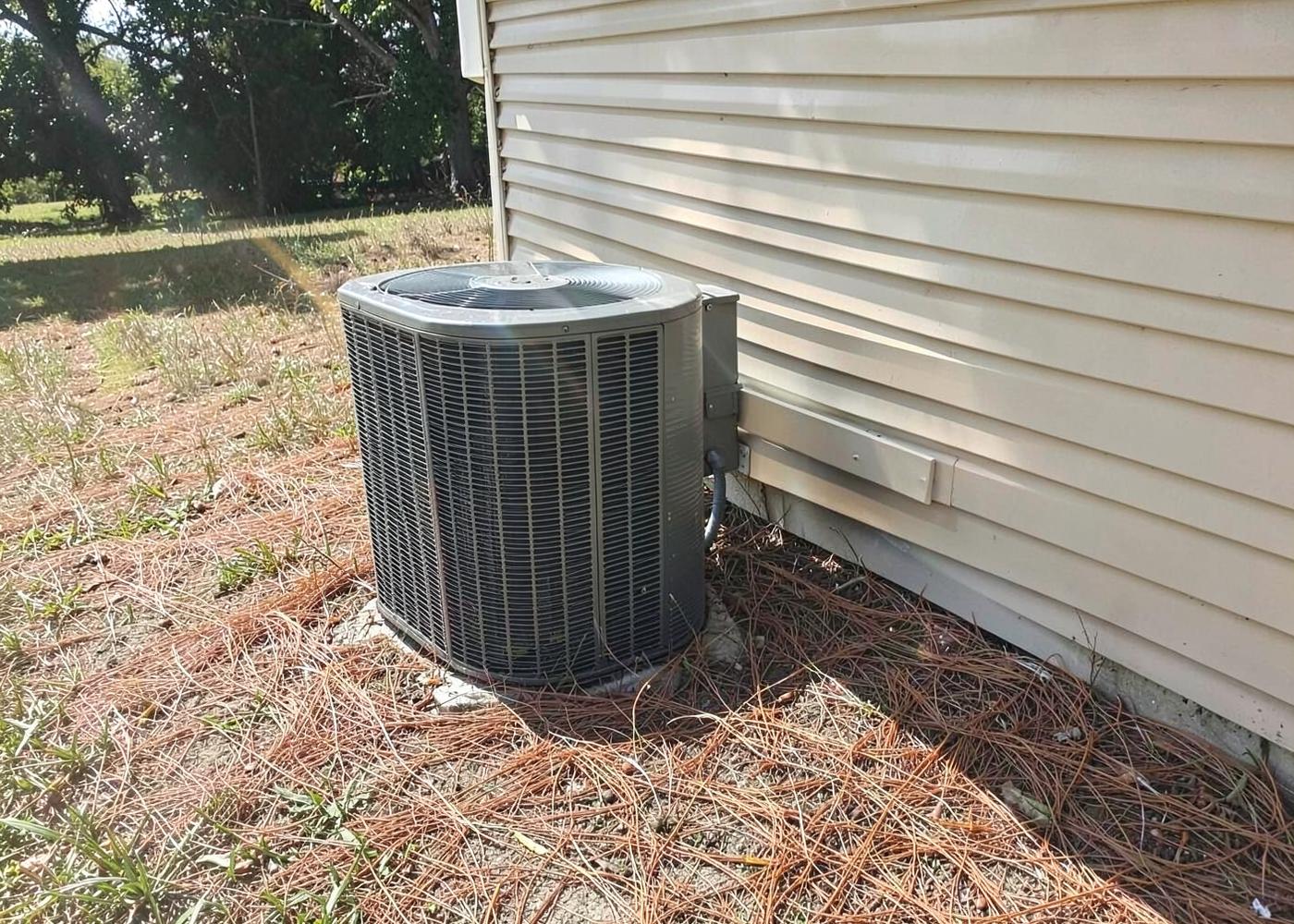 Outdoor air conditioning condenser unit running beside a brick house wall in bright hazy heat with overgrown grass around the pad