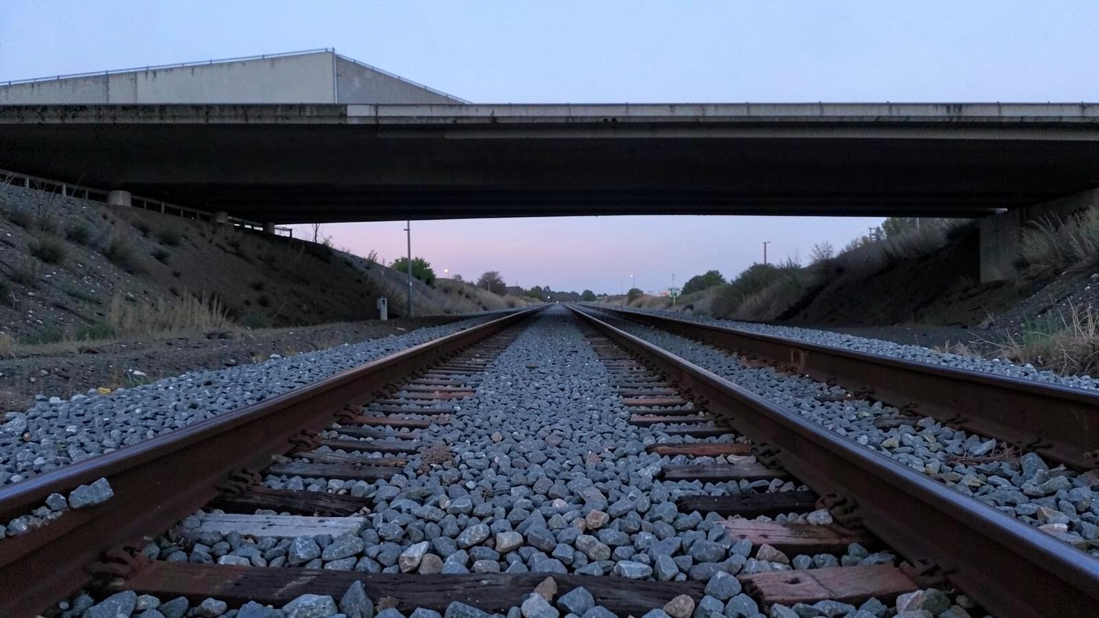 Low view along converging steel rail lines on gravel ballast beneath a plain concrete highway overpass at dusk