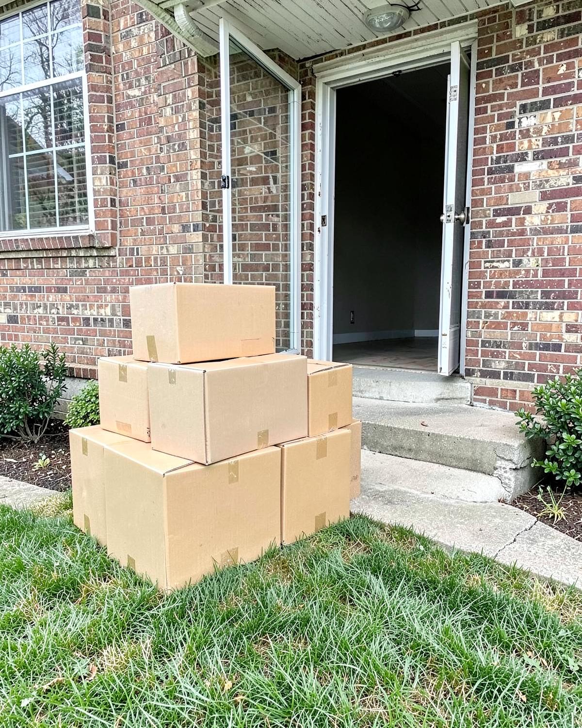 Plain cardboard moving boxes stacked on the lawn beside the concrete steps of a brick house with the front door open to an empty room