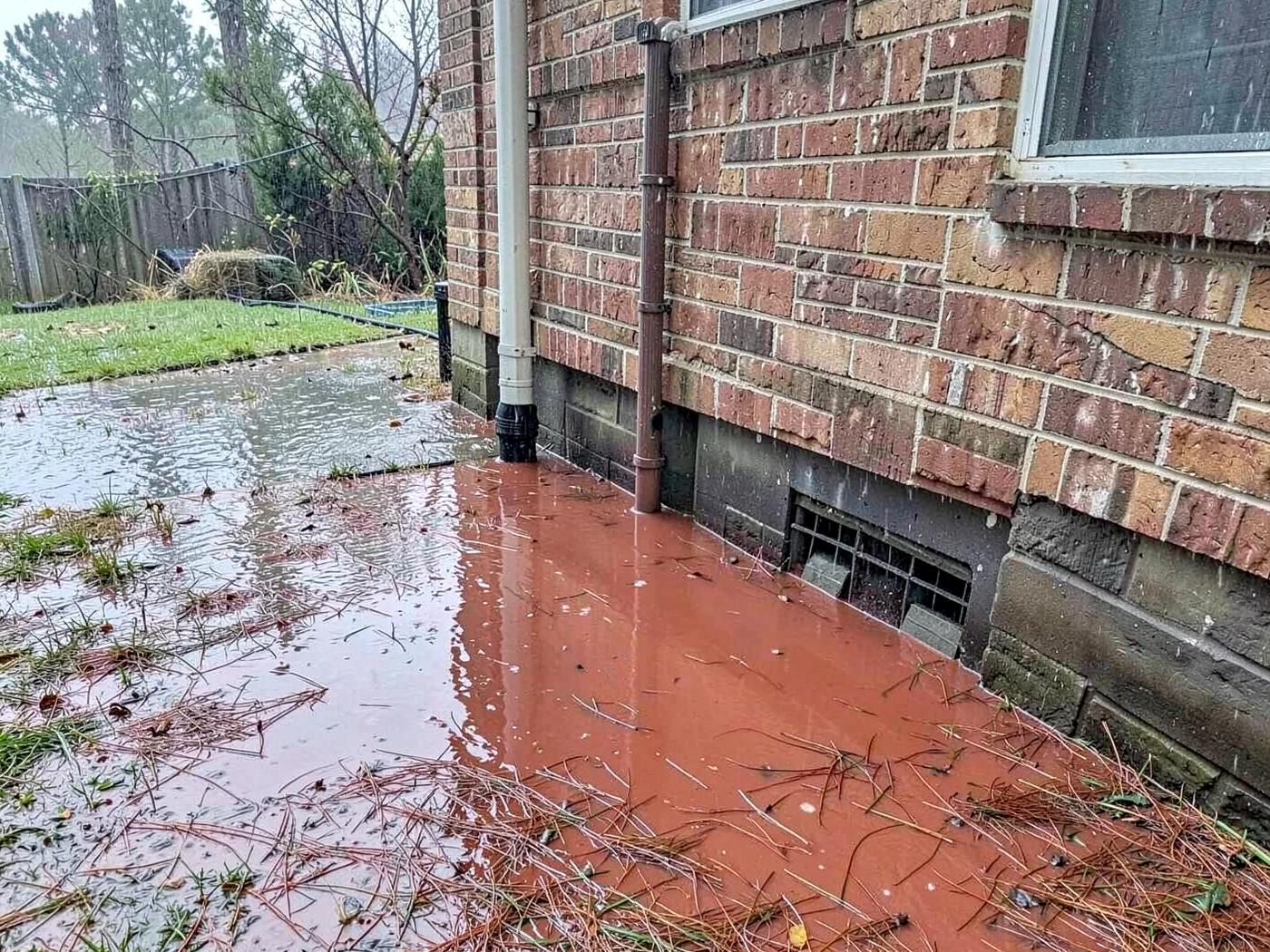 Muddy red rainwater pooling against the brick foundation and crawlspace vent of a house after a heavy summer thunderstorm