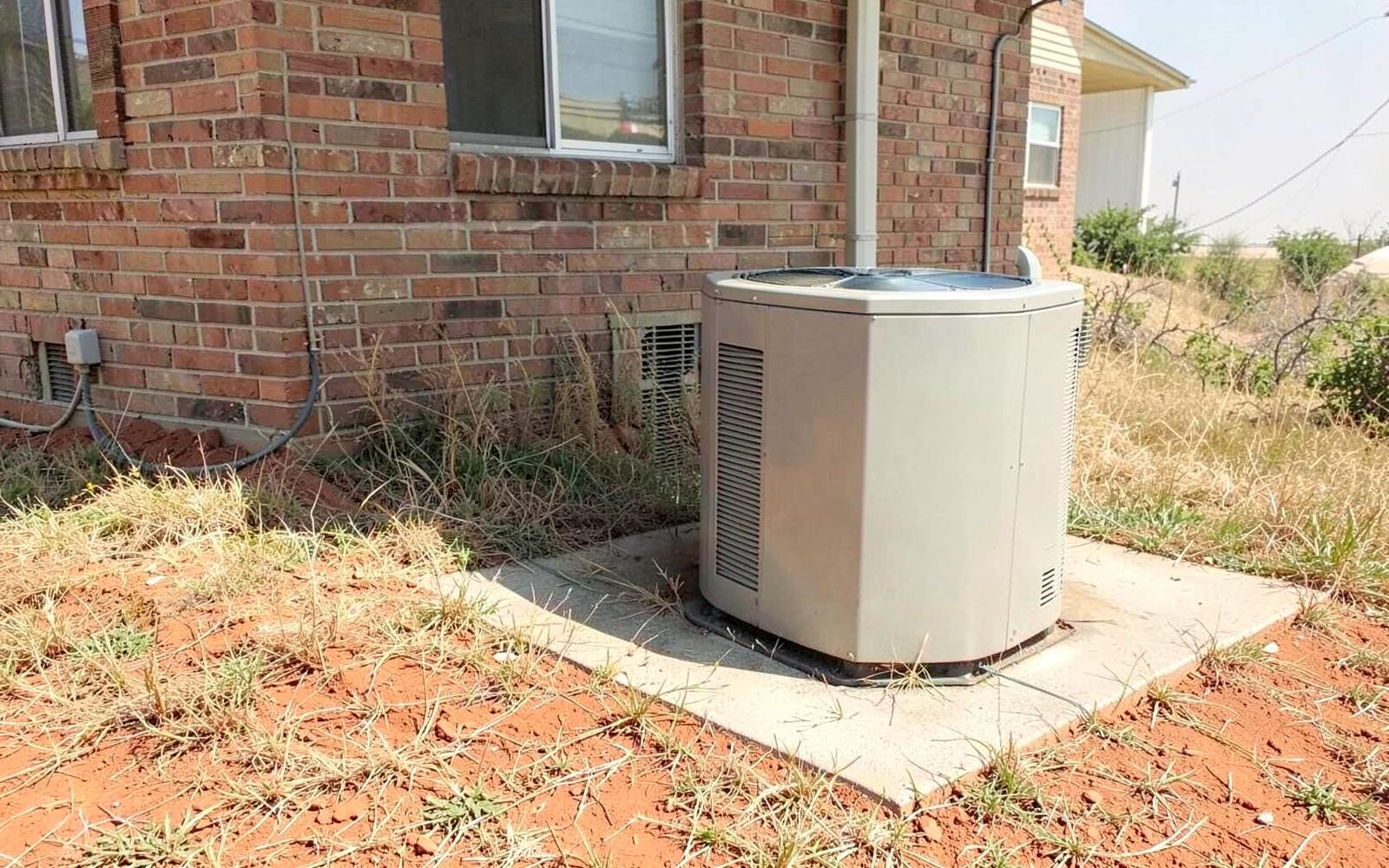 Plain outdoor air conditioning condenser on a concrete pad beside a brick house in hazy summer heat with dry grass and red clay soil