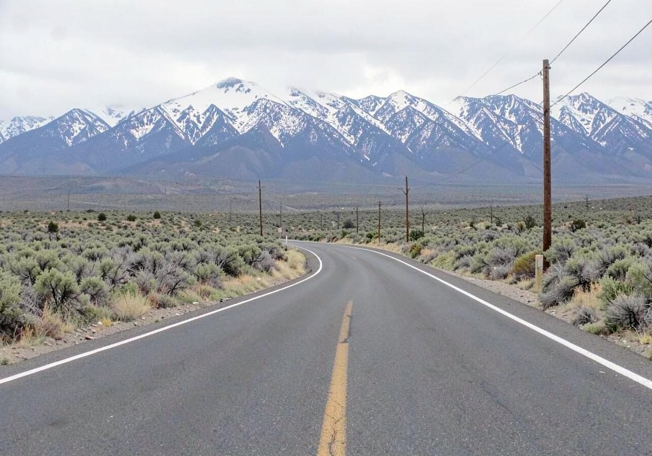 Empty two-lane highway curving through sagebrush country toward snow-covered mountains under an overcast sky