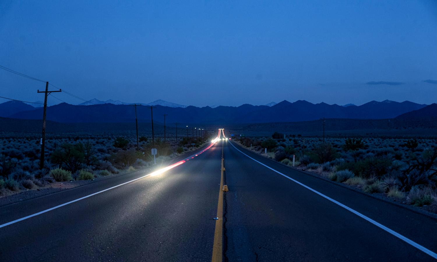 Two-lane highway crossing an open sagebrush basin at blue hour with headlight streaks and a silhouetted mountain range