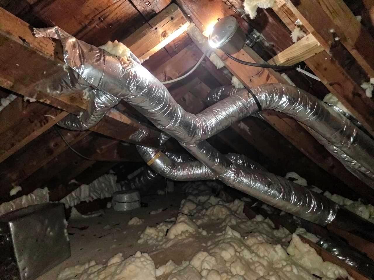Flexible ductwork running across joists in a hot unfinished attic lit by a single work light