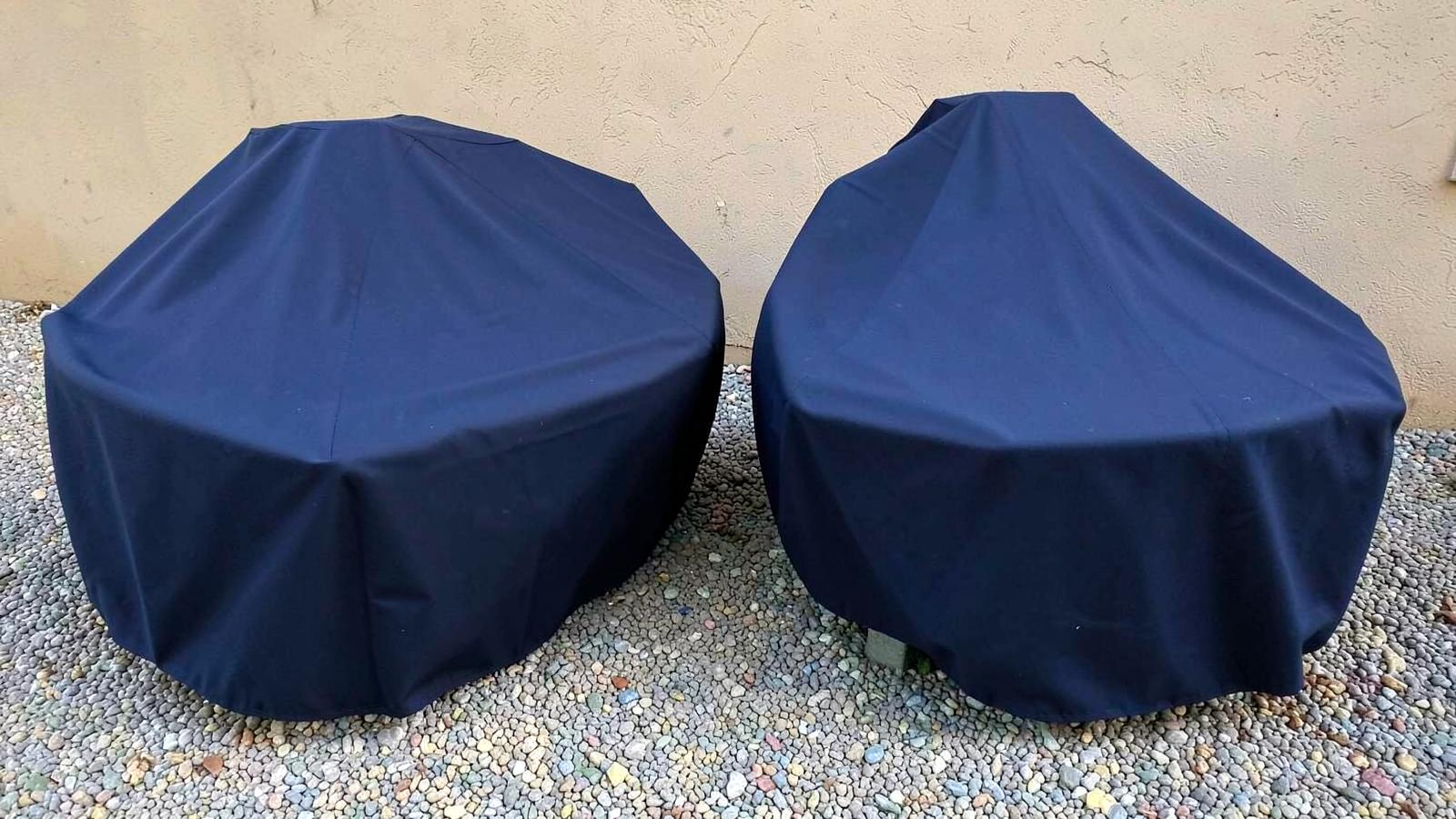 Two boats fully wrapped in plain navy covers parked on gravel beside a stucco wall in bright sun