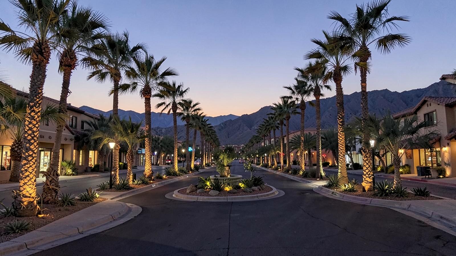 Palm-lined upscale desert boulevard at dusk with landscape lighting, a planted roundabout and mountain silhouette