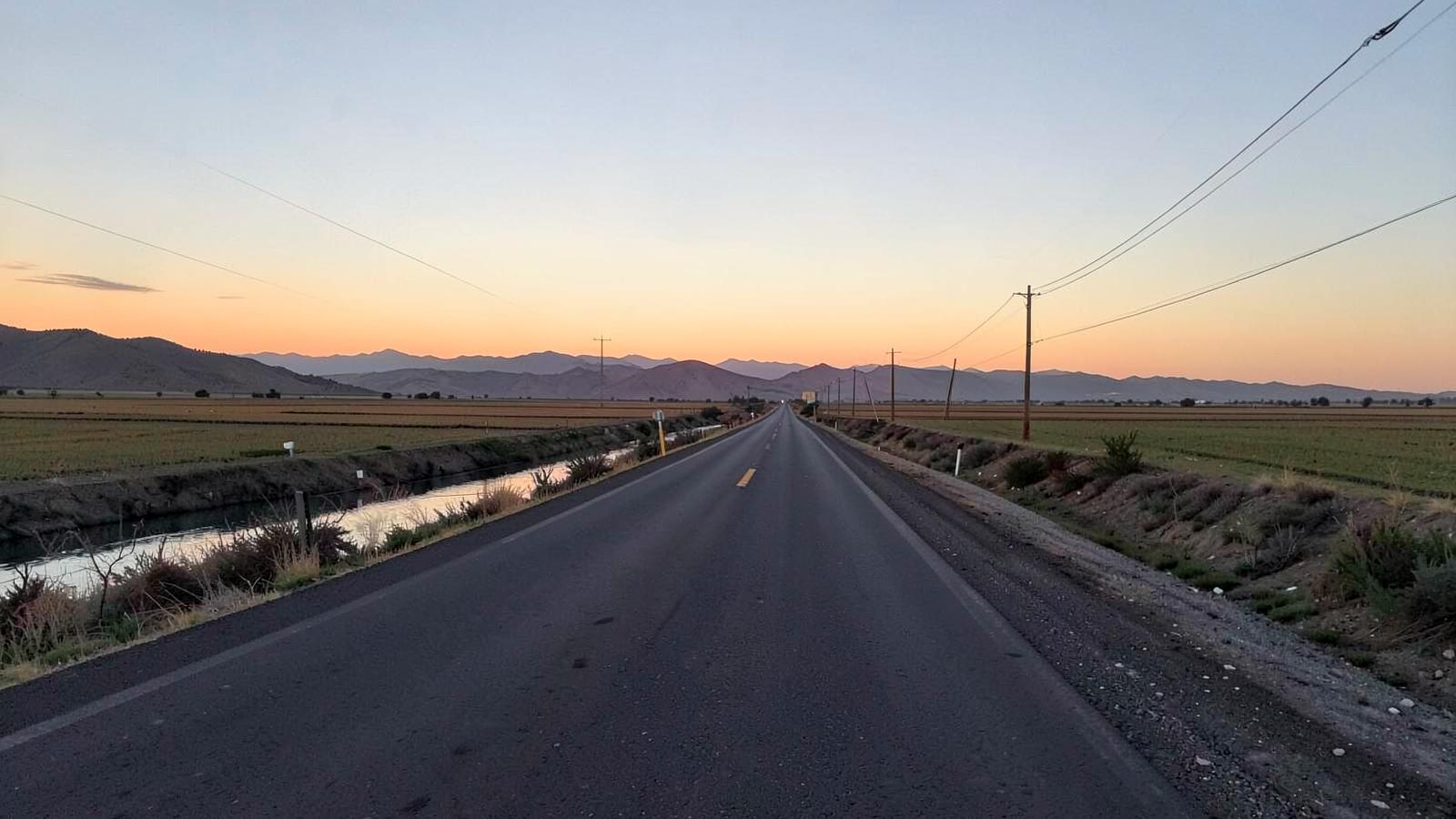 Empty straight desert road crossing flat farmland at sunrise with an irrigation canal alongside and arid ridges beyond