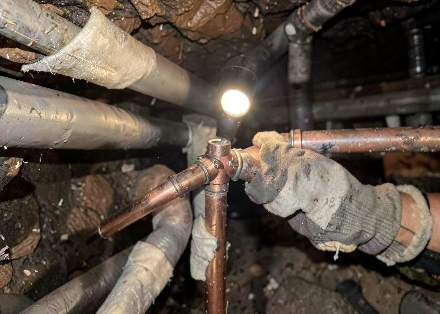Gloved hands fitting a copper pipe joint among insulated lines in a mountain home crawlspace lit by a hand lamp