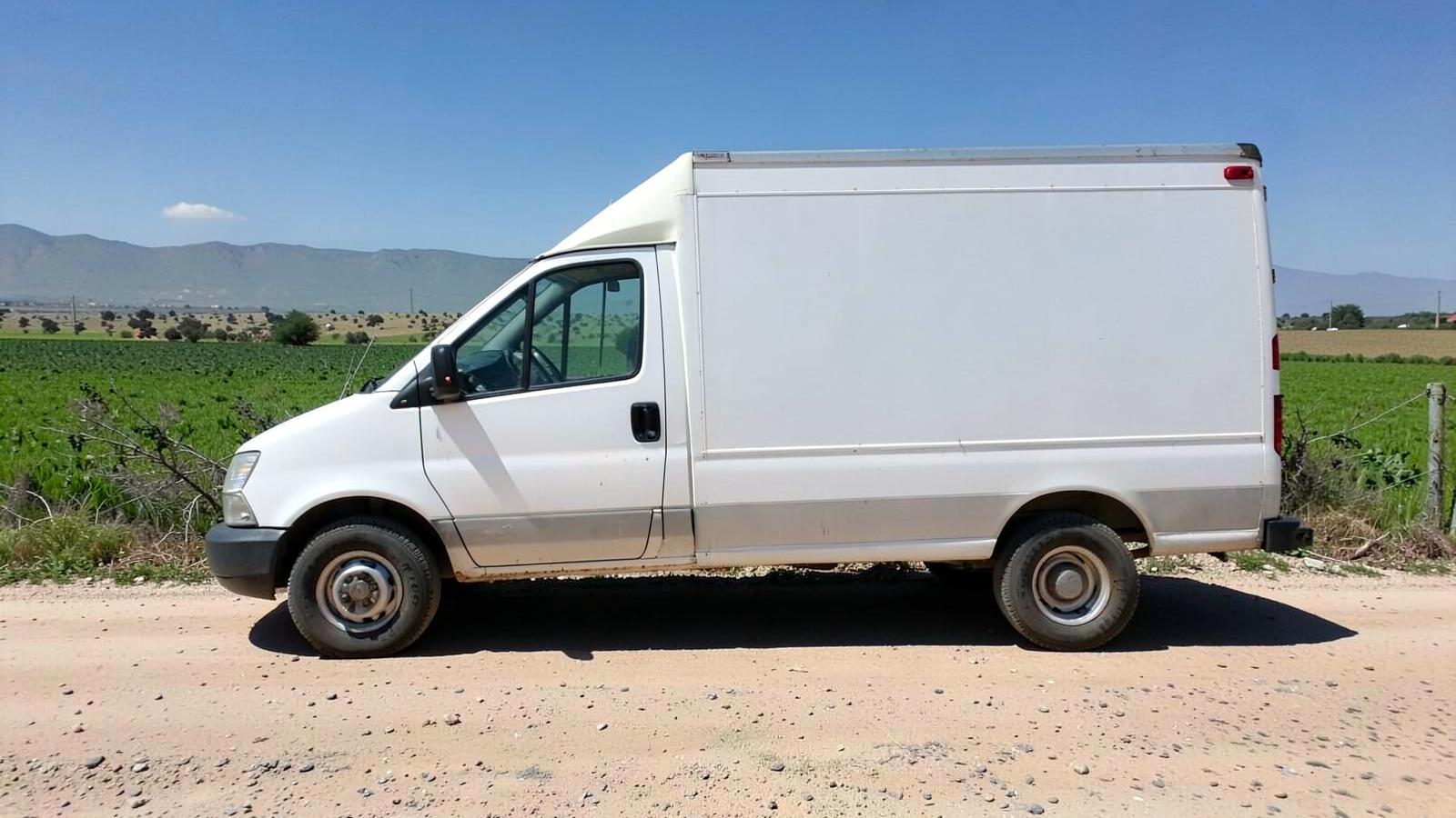 Plain unmarked white cargo work van parked on a dusty farm road beside green desert fields in harsh sunlight