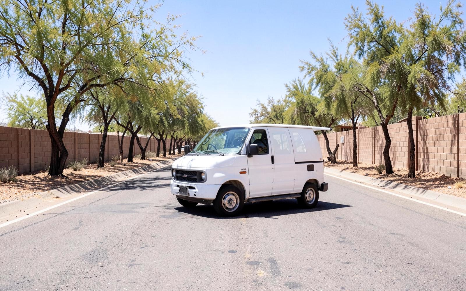 Plain white service van parked on a wide sunbaked desert arterial road lined with palo verde trees and block walls