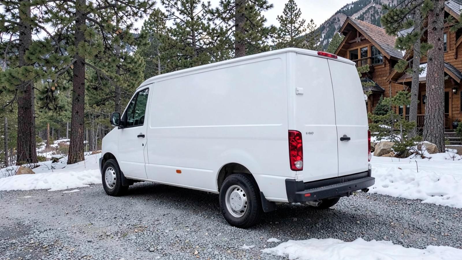 Plain white cargo van with blank panels parked on a snowy gravel driveway beside pines and a timber mountain home