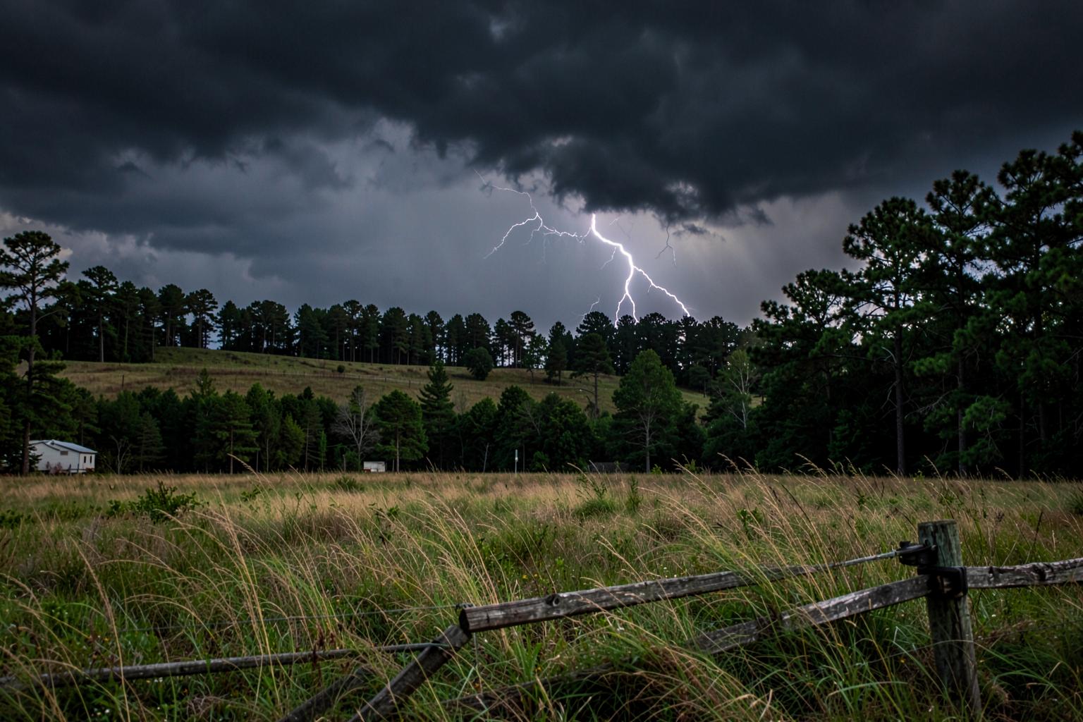 Dark thunderstorm and lightning building over open pasture and a pine treeline behind a wooden fence