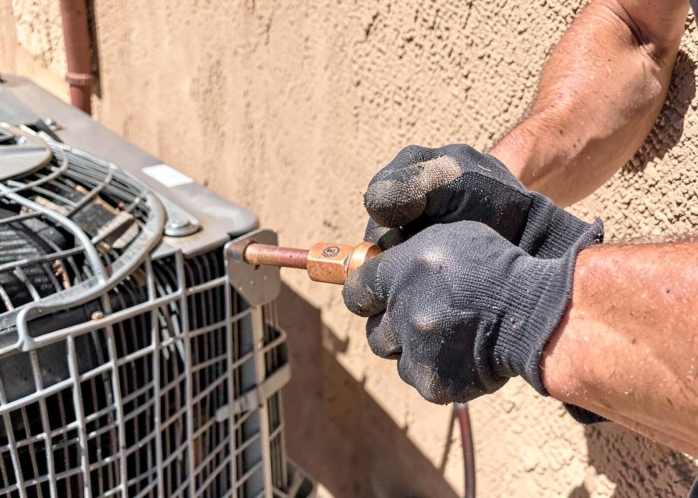 Close-up of a technician's gloved hands fitting a copper line to an air conditioning condenser beside a stucco wall in harsh sun