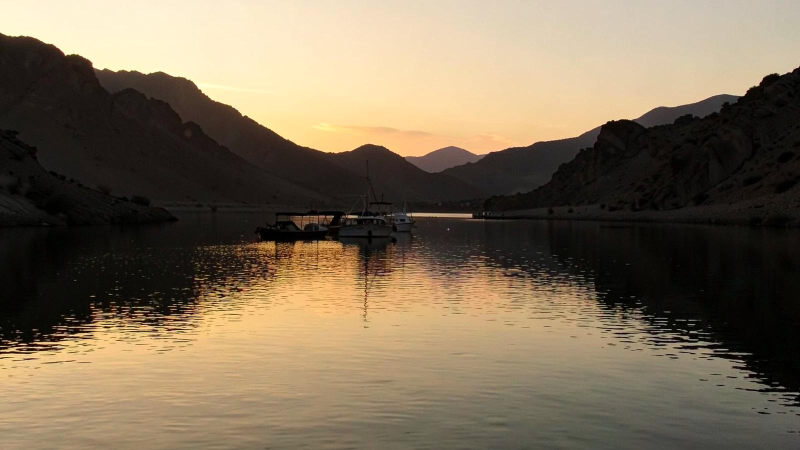Calm desert lake channel at dusk with moored boats in silhouette and arid ridges under a warm orange sky