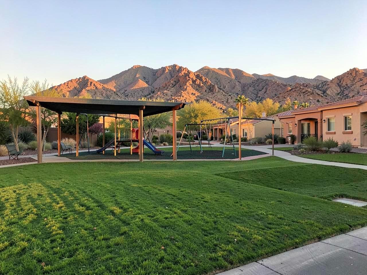 Suburban neighborhood park with a shaded playground and green grass below desert mountains at golden hour