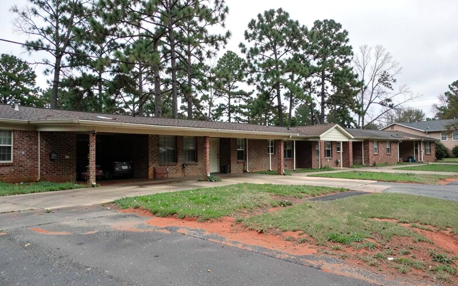 Quiet street of single-story mid-century red brick ranch homes with carports and tall pines along the verge