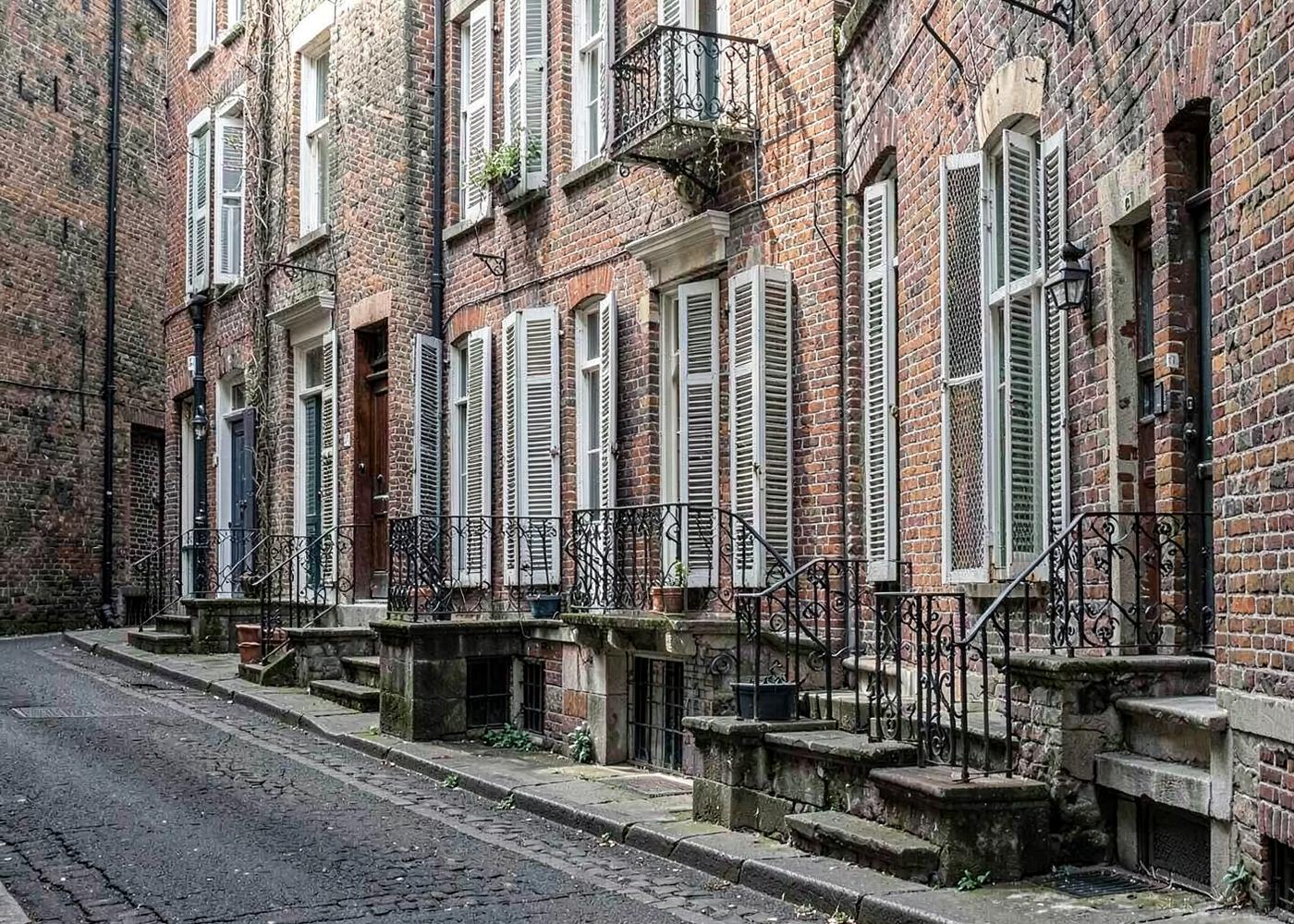 Row of nineteenth century brick townhouses with tall shuttered windows and cast iron stair railings along a narrow street