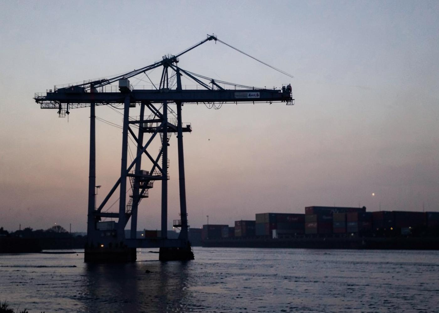 Container terminal gantry crane silhouetted above a wide river channel at dusk with stacked containers in the distance
