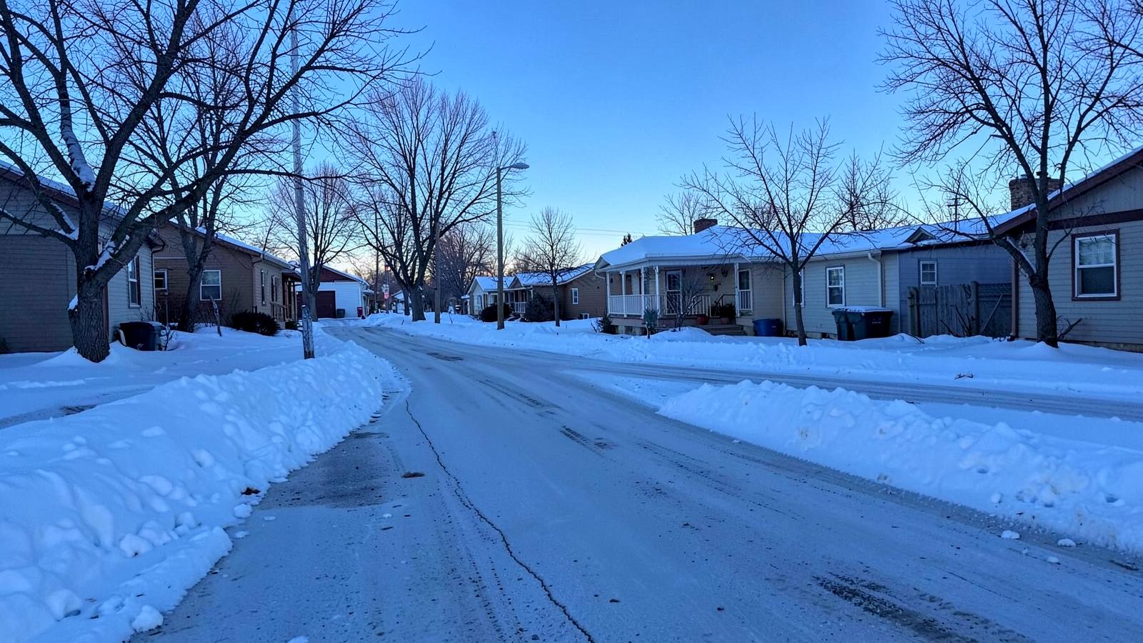 Snow-covered residential street with modest single-story houses, bare trees and deep snowbanks at the curb