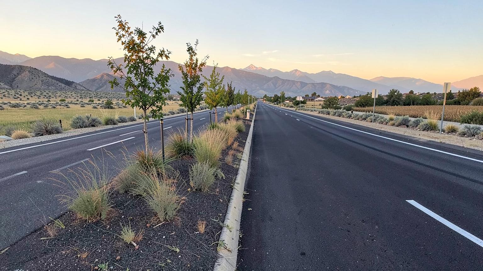 Wide new suburban arterial road with landscaped median and young trees running past open farmland toward mountains