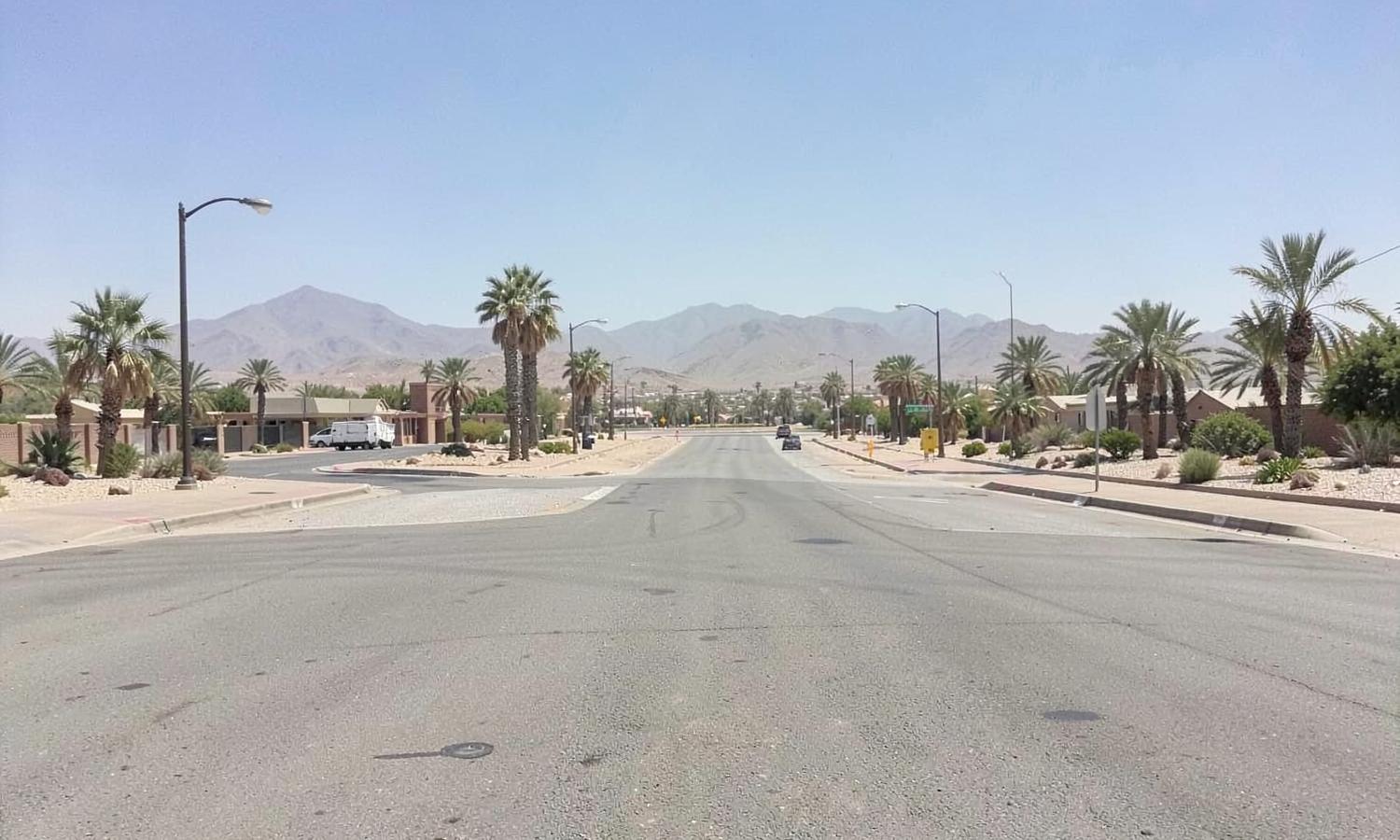 Wide flat desert boulevard with palm trees and gravel medians under midday sun with distant mountain ranges