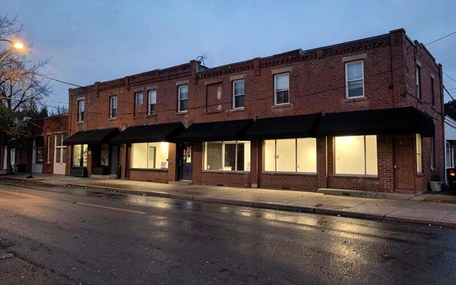 Row of plain two-story red brick small-town buildings at dusk with dark awnings and wet reflective pavement