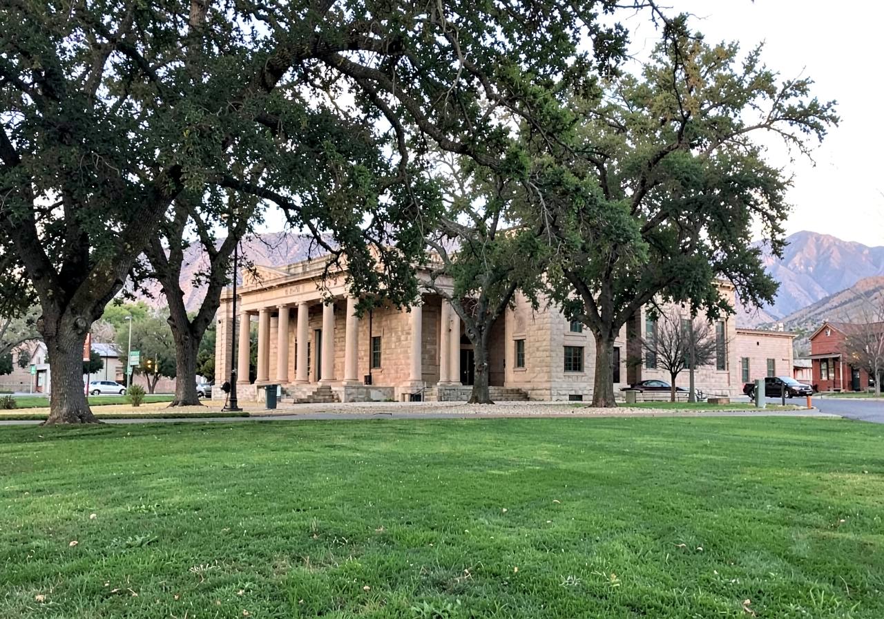 Sandstone classical building with columns behind mature shade trees on a wide green lawn in a small western town