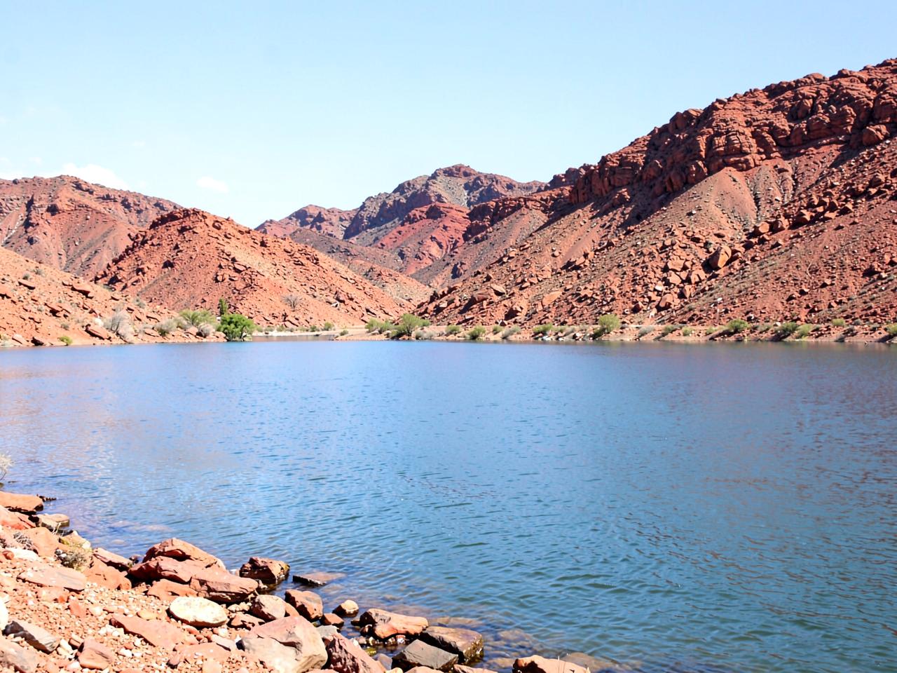 Desert lake shoreline with rocky red-brown hills and calm blue water under bright sky
