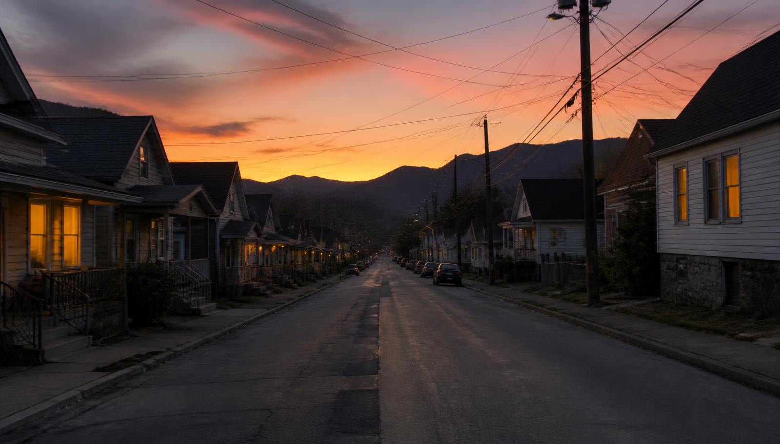Evening view down a wide residential street of closely spaced modest homes with lit windows and mountains silhouetted