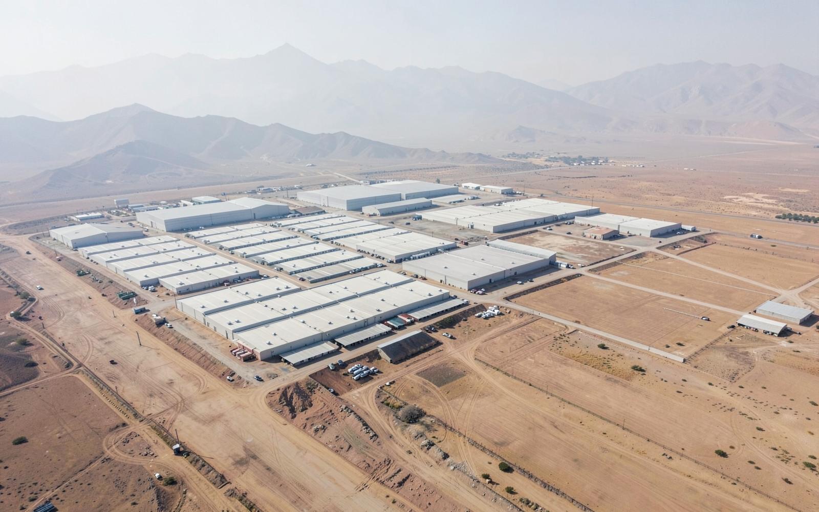 Aerial view of a desert valley edge where a grid of new rooftops meets large blank-roofed warehouses and open land