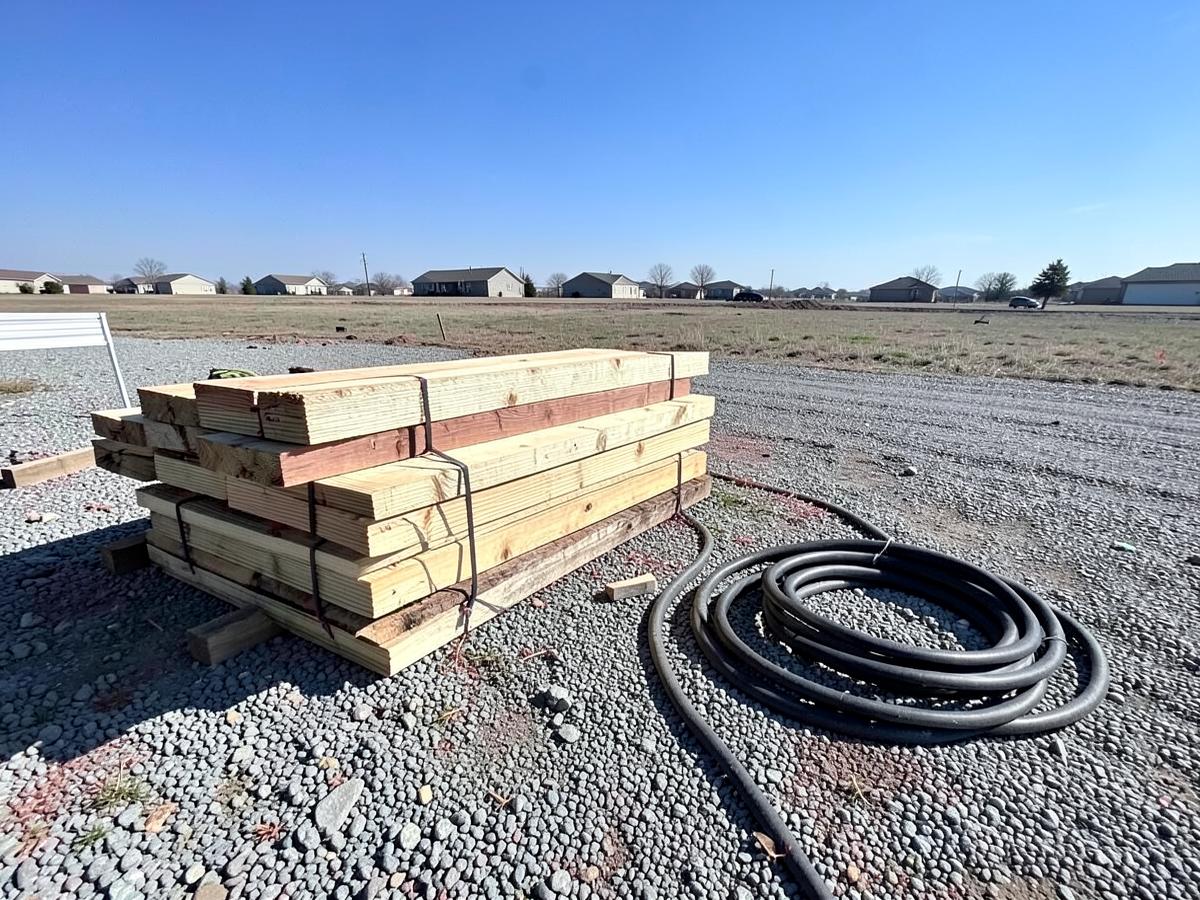 Stacked fresh lumber and a coiled hose on gravel at a residential jobsite on a wide flat lot