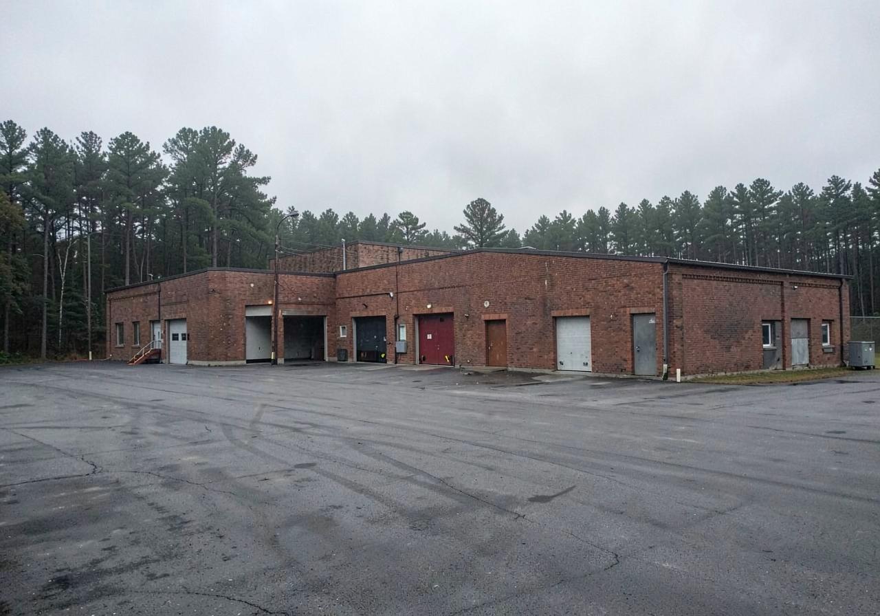 Brick warehouse with loading bays and an empty paved yard backed by a pine treeline on an overcast morning