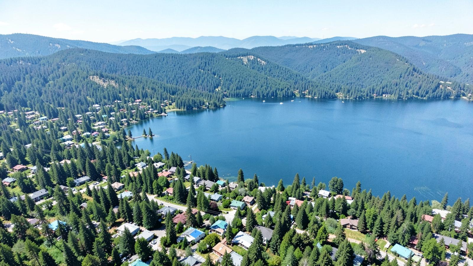 Aerial view of a forested lakeshore town with evergreen hills meeting a broad blue lake