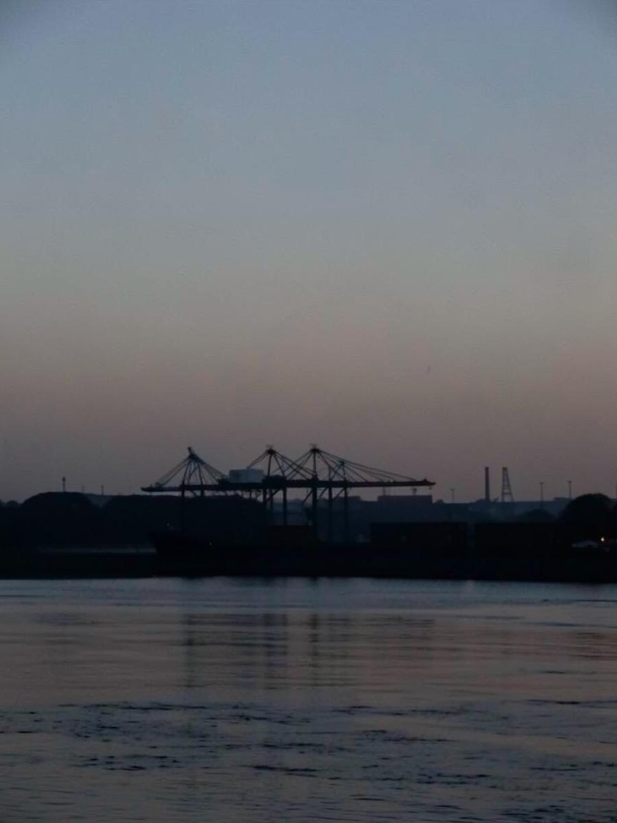 Silhouetted container port cranes against a hazy dusk sky across a wide coastal river