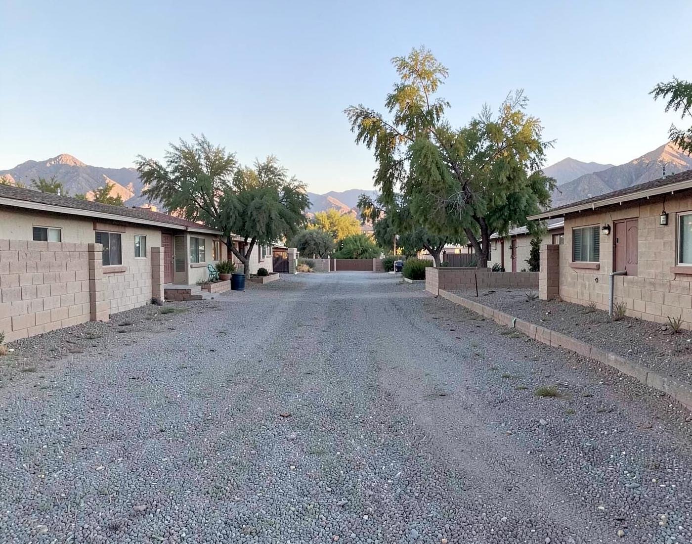 Quiet street of modest single-story desert homes with low block walls, gravel lots and mature shade trees