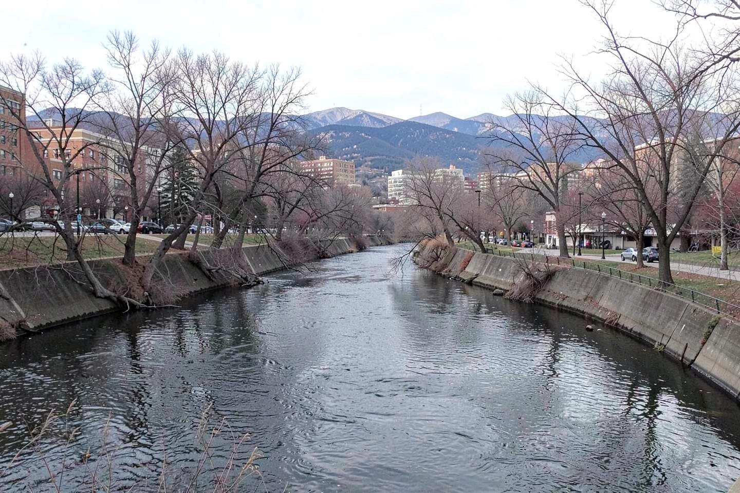River running through a downtown park with bare winter trees and mountains rising in the distance