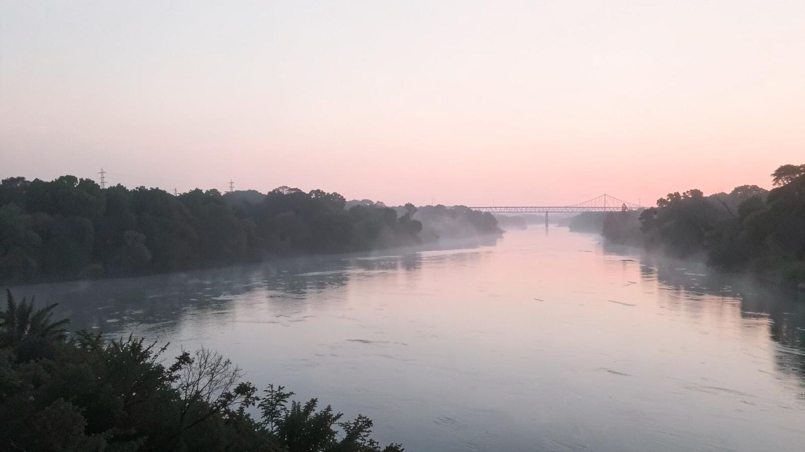 Dawn mist rising off a wide slow inland river with wooded banks and a distant steel trestle silhouette in soft pink light