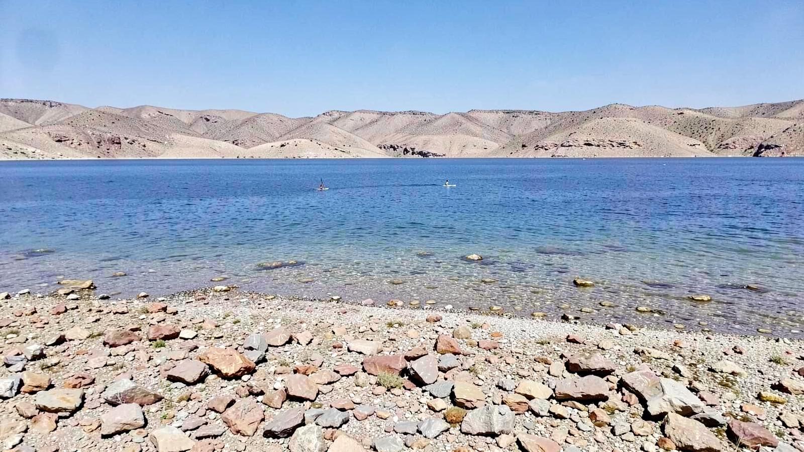 Rocky desert lake shoreline with clear blue water, a distant paddleboarder and bare arid ridges beyond