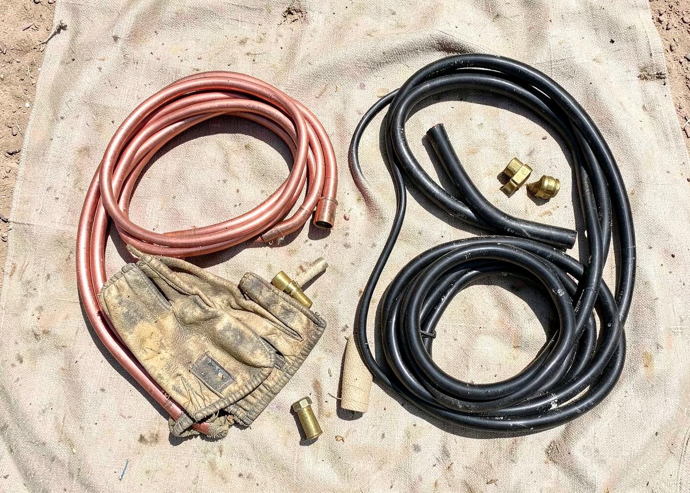 Overhead view of a coil of copper pipe, brass fittings, worn leather gloves and black tubing on a dusty canvas tarp in desert sun